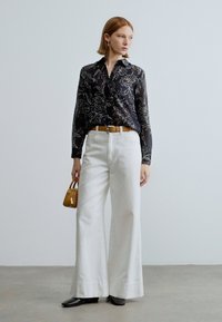 Camisa preta com padrão floral, abotoada, de mangas compridas, combinada com calças brancas de perna larga e cinto castanho. O modelo segura uma pequena mala castanha.