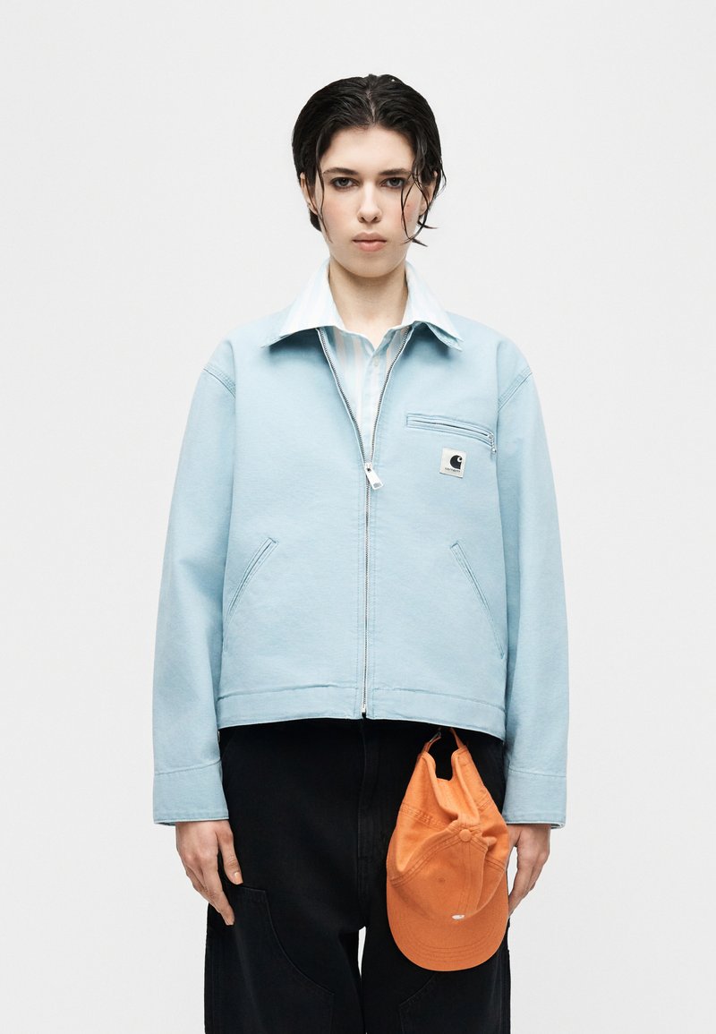 Jeune personne aux cheveux courts noirs mouillés portant une veste zippée bleu clair, un pantalon noir, tenant une casquette orange à côté d'elle.