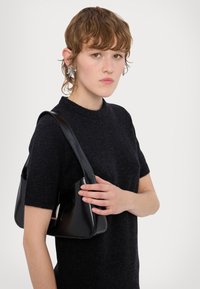 Svart patentlæder håndtaske med en buet, struktureret form, båret over skulderen. Modellen har en mørk kortærmet strikket bluse på.