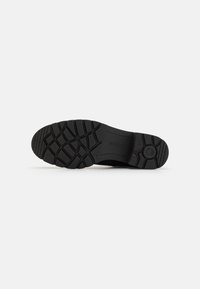 Semelle de chaussure en caoutchouc noir avec un motif de semelle texturé, comportant un détail circulaire central et le logo de la marque. Design plat pour la stabilité.