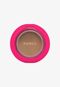 Compact rond en silicone rose vif avec un couvercle transparent révélant un produit brun clair à l'intérieur, embossé avec "FOREO" en or.