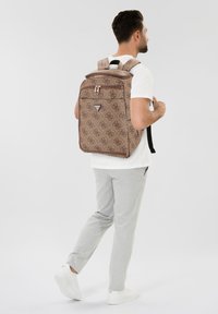 Rucsac bej cu model, dotat cu două fermoare, bretele reglabile și un buzunar frontal. Material texturat cu un design repetitiv al logo-ului.