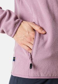Fleece-takki vaaleanliilana, jossa on vetoketjullinen tasku, sileä pinta ja kontrastivärit hemman ja hihansuiden reunassa.