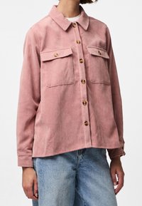 Vrouw in een roze corduroy overhemddoek met twee borstzakken en lichtblauwe denim jeans.