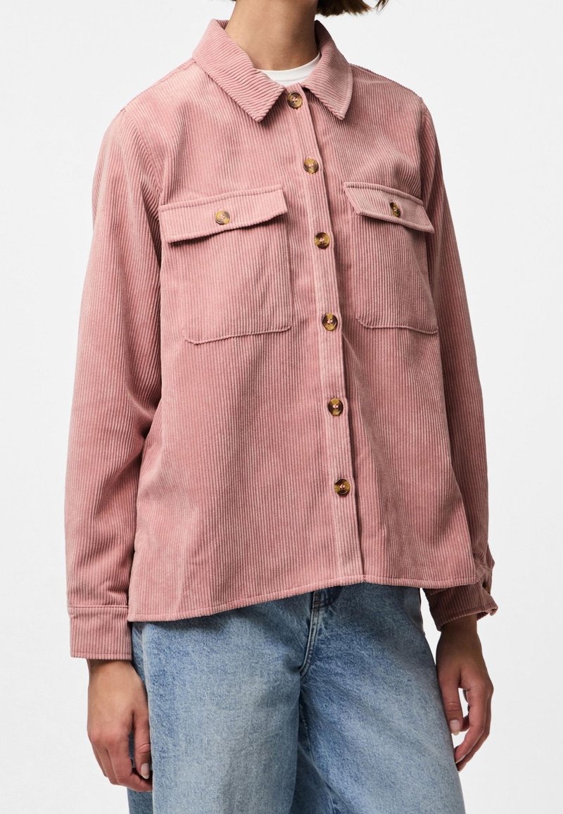 Vrouw in een roze corduroy overhemddoek met twee borstzakken en lichtblauwe denim jeans.