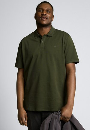 Jeune homme portant un polo vert foncé, tenant une veste grise pliée dans sa main droite sur un fond gris uni.