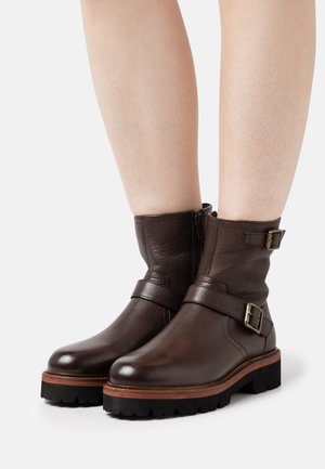 Botas de tornozelo em couro castanho com detalhes em fivela, material texturizado e solas de borracha. Apresenta um fecho lateral e uma entressola castanha em contraste.