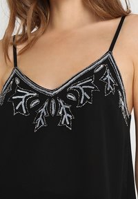 Camisole noire avec un décolleté en V, ornée de motifs floraux argentés brodés, avec des bretelles fines et réglables, et une texture de tissu léger.