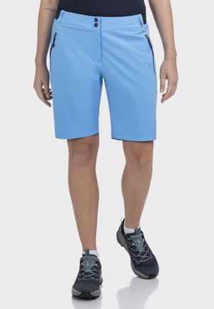 Person trägt hellblaue Kniebundhose mit Reißverschlusstaschen vorne und schwarze sowie graue Sportschuhe und steht vor weißem Hintergrund.