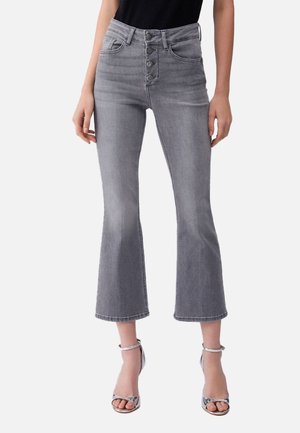 Donna che indossa jeans grigi a vita alta, taglio svasato e corti, con chiusura a bottoni, abbinati a un top nero e sandali argentati con tacco alto.