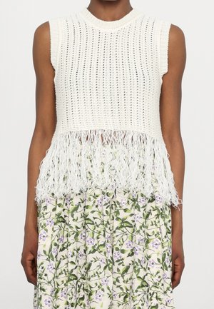Haut blanc en crochet sans manches avec ourlet à franges, porté sur une jupe crème avec un motif de feuilles vertes et de petites fleurs violettes.