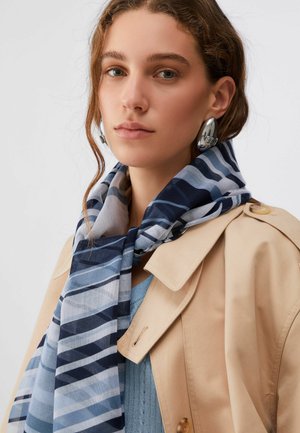 Giovane donna che indossa orecchini a cerchio d'argento, un trench beige, un maglione azzurro e una sciarpa a righe blu-grigio intorno al collo.