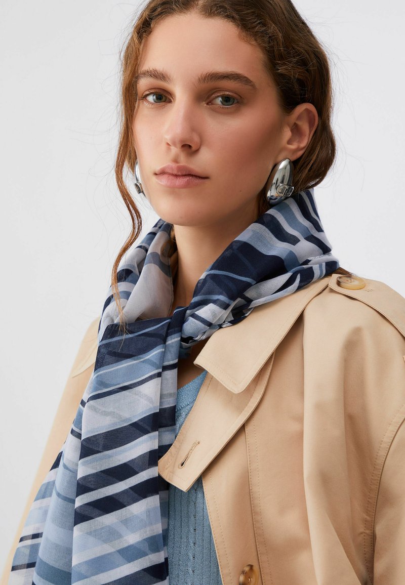 Jeune femme portant des boucles d'oreilles créoles argentées, un trench-coat beige, un pull bleu et une écharpe rayée bleu-gris autour du cou.