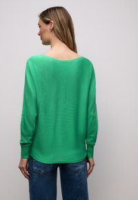 Femme vue de dos portant un pull vert vif à manches longues et un jean bleu sur un fond gris clair uni.