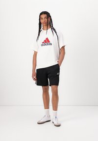 Hvid t-shirt med rødt og marineblåt Adidas-logo, sorte shorts og hvide sneakers med beige accenter, stående foran en ensfarvet baggrund.