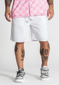 Pantaloni corti bianchi con elastico in vita, indossati con sneakers grigie e nere. Tatuaggi visibili sulle gambe. Lo sfondo è semplice e di colore chiaro.
