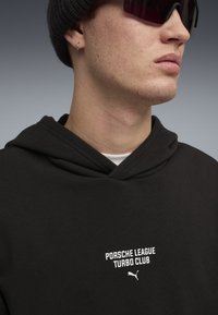 Čierna mikina s kapucňou obsahuje biely nápis "PORSHE LEAGUE TURBO CLUB" a logom Puma. Model má na hlave čiapku a tmavé slnečné okuliare.
