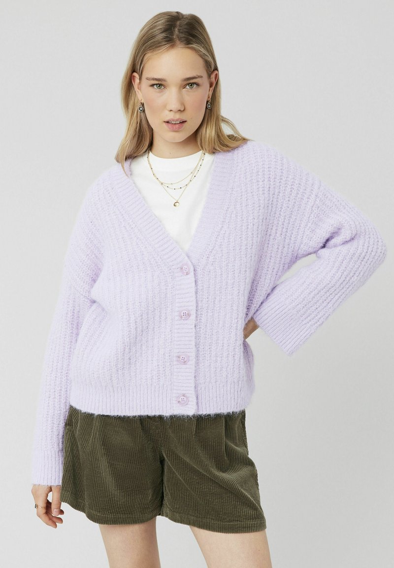 Cardigan tricoté lavande avec un col en V, fermeture à boutons et texture côtelée. Associé à un short en velours côtelé vert olive.