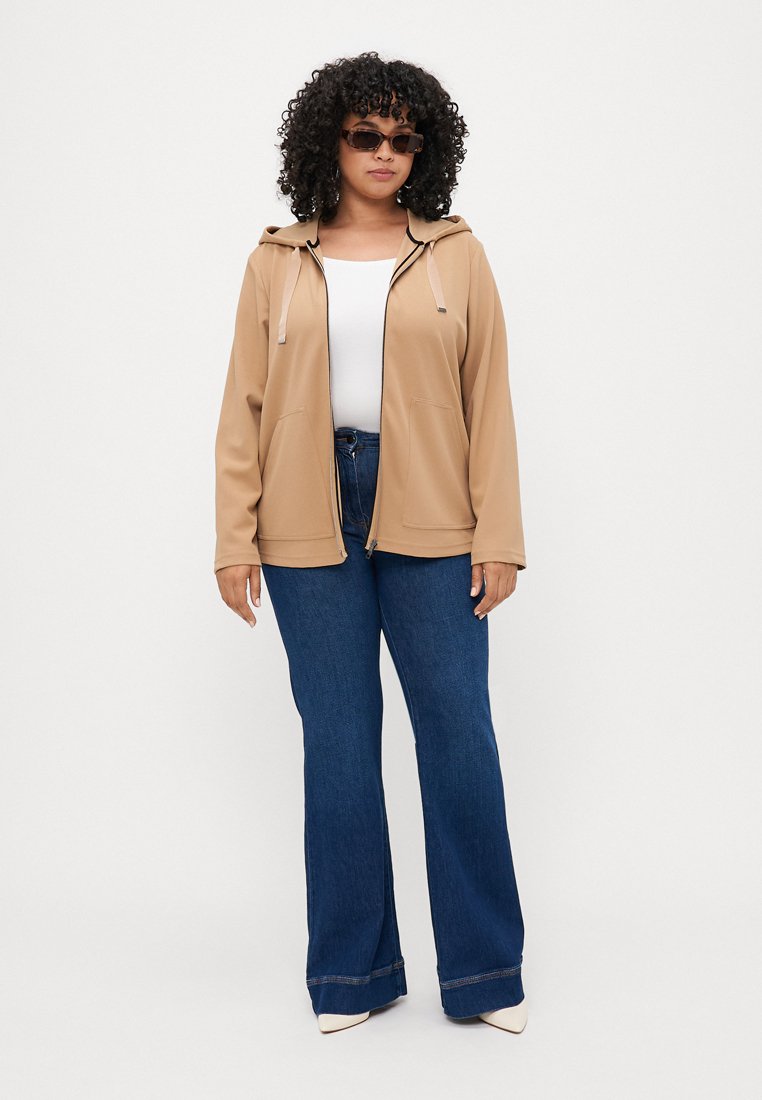 Veste à capuche beige avec poches avant, associée à un jean évasé bleu. Le modèle porte un haut blanc et des lunettes de soleil foncées.