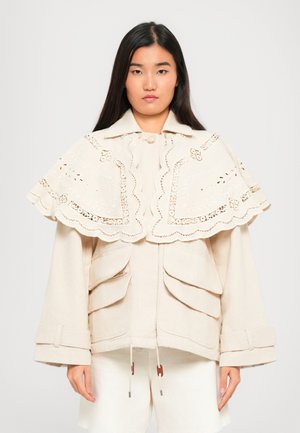 Jonge vrouw met lang, donker haar, gekleed in een beige jas met grote voorzakken en een geborduurde kantenkraag.
