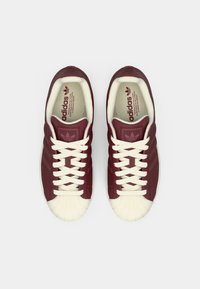 Μπορντό δερμάτινα αθλητικά παπούτσια με λευκά κορδόνια, υφασμένο καουτσούκ στο πάνω μέρος των δακτύλων και σήμανση Adidas στη γλώσσα και στην εσωτερική σόλα, με κλασικό σχέδιο.