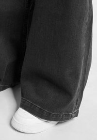 Jeans anchos negros de mezclilla, con un dobladillo sin acabado y costuras visibles, combinados con zapatillas blancas en la parte inferior.