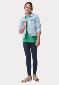 Giacca di jeans azzurro chiaro sopra una maglietta verde, abbinata a leggings navy e sandali dorati con cinturini, che mettono in mostra un outfit casual.
