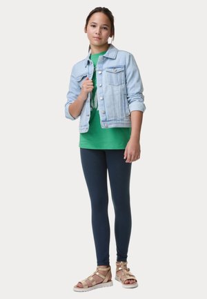 Veste en denim bleu clair sur un t-shirt vert, associée à des leggings navy et des sandales dorées à lanières, illustrant une tenue décontractée.
