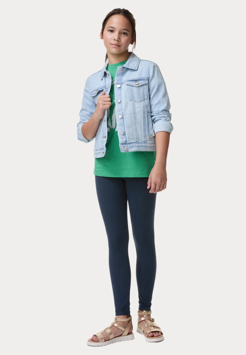 Giacca di jeans azzurro chiaro sopra una maglietta verde, abbinata a leggings navy e sandali dorati con cinturini, che mettono in mostra un outfit casual.