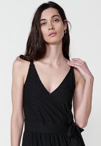 Schwarzes Wickelkleid mit V-Ausschnitt, dünnen Trägern, strukturiertem Stoff und tailliertem Detail. Verfügt über ein dezentes geometrisches Muster.