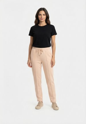 Femme debout portant une chemise noire à manches courtes, un pantalon beige à cordon de serrage et des baskets beiges sur un fond blanc uni.