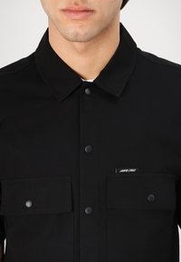 Čierna košeľa s gombíkmi vyrobená z ľahkého materiálu, s špicatým golierom, dvoma náprsníkovými vreckami a malou logom na ľavom vrecku.