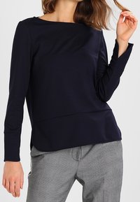 Tmavě modrý top s dlouhým rukávem a lodním výstřihem, těsně přiléhavý střih a boční rozparky, kombinovaný se šedými károvanými kalhotami. Hladká textura tkaniny.