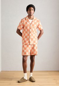 Rövid ujjú ing és rövidnadrág szett narancssárga és fehér geometrikus mintával, gombos záródás, gesztenyebarna slip-on cipővel és fehér zoknival párosítva.