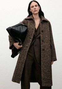 Abrigo de lana texturizado marrón con cuello tipo solapa, cierre de botones y bolsillos laterales, llevado sobre un traje marrón oscuro, portando un bolso de cuero negro.