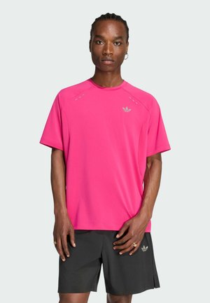 Uomo con capelli intrecciati che indossa una maglietta Adidas rosa acceso e pantaloncini neri, in piedi su uno sfondo grigio chiaro uniforme.