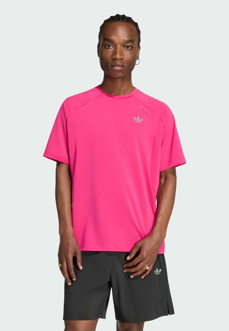 Hombre con trenzas, que lleva una camiseta rosa brillante de Adidas y pantalones cortos negros, de pie frente a un fondo liso de color gris claro.