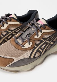 Sport sneakers with a mix of brown and beige mesh fabric, textured suede accents, gray overlays, and black laces. Pink branding on the tongue.