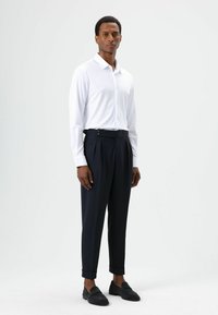 Wit shirt met lange mouwen en knoopsluiting, gecombineerd met marineblauwe, hooggetailleerde broek met plooien en opgerolde boorden. Zwarte instapschoenen.