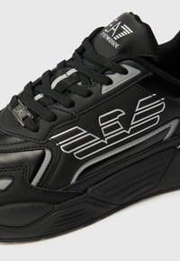 Sorte joggesko med et stilrent design laget av syntetisk lær, teksturerte detaljer, sølvfarget logo og kontrasterende svarte snører.