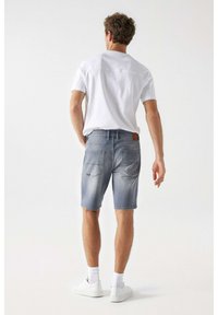 Pantalones cortos de mezclilla azul claro con un ajuste relajado, que cuentan con bolsillos traseros y un parche de cuero marrón, combinados con una camiseta blanca lisa y zapatillas blancas.