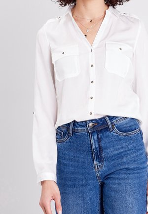 Femme portant une chemise blanche à boutons avec poches poitrine et un jean bleu taille haute, visible du cou aux hanches sur un fond uni.