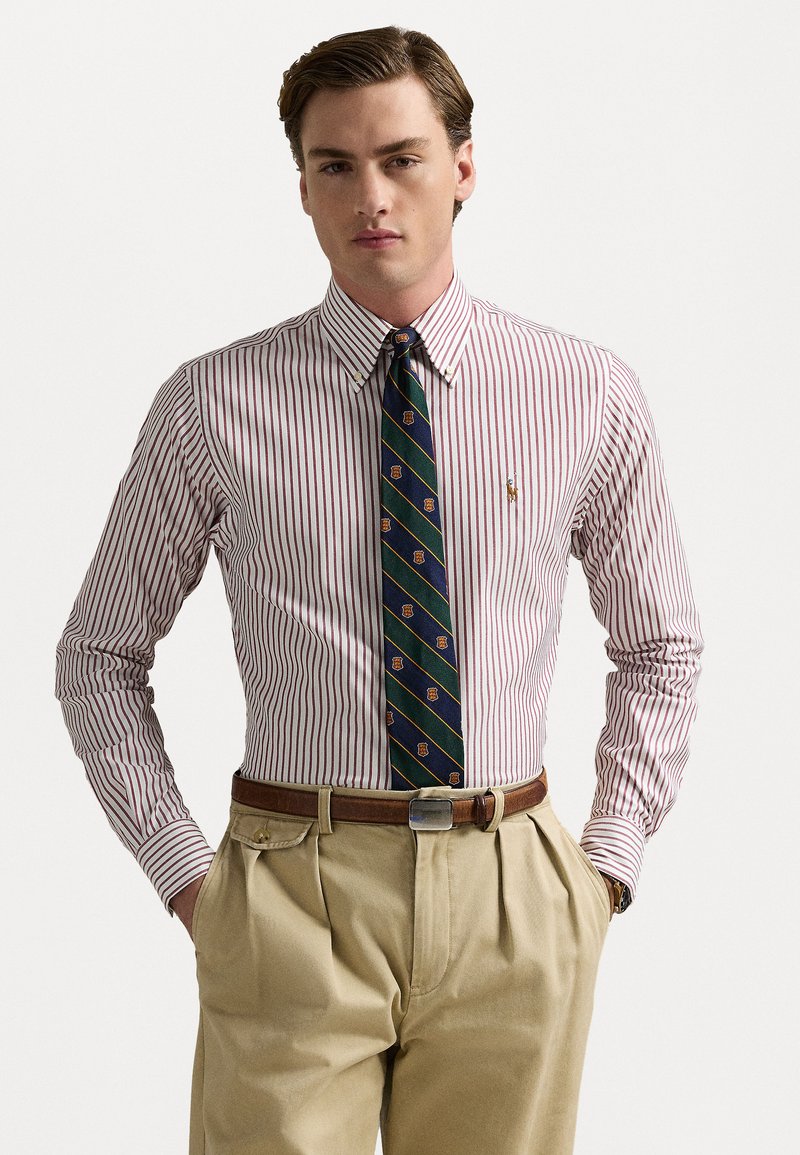 Chemise blanche à rayures verticales rouges, col boutonné, et une cravate foncée avec un motif géométrique. Pantalons beiges avec une ceinture marron.