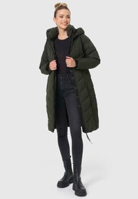 Langer, dunkelgrüner Daunenmantel mit Kapuze, gestepptem Design, Reißverschluss auf der Vorderseite und elastischen Bündchen, kombiniert mit schwarzen Skinny-Jeans und klobigen schwarzen Stiefeln.