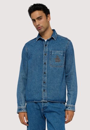 Camisa de denim azul con un bolsillo frontal y diseño bordado, con cierre de botones y mangas largas. Textura suave.