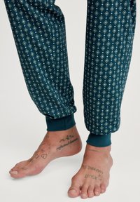 Katoenen pyjamabroek in donker teal met een ruitpatroon in lichtblauw en groen. Geribbelde boorden bij de enkels; zichtbare blote voeten met tatoeages.