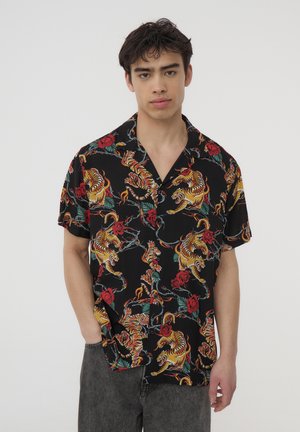 Giovane uomo con una camicia nera a maniche corte con stampa colorata di tigre e rosa, abbinata a pantaloni grigio scuro, in piedi contro uno sfondo bianco.