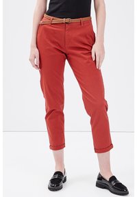 Personne portant un pantalon rouille à mi-mollet avec des ourlets retroussés, une ceinture tressée marron, un haut noir et des mocassins noirs en vernis, se tenant sur un sol blanc.