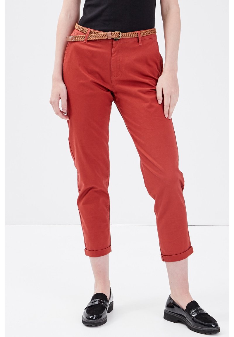 Personne portant un pantalon rouille à mi-mollet avec des ourlets retroussés, une ceinture tressée marron, un haut noir et des mocassins noirs en vernis, se tenant sur un sol blanc.