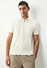 Beige polo met korte mouwen, gemaakt van zacht materiaal, met een kraag met twee knopen en een klein geborduurd logo op de borst. Draag het met beige shorts.